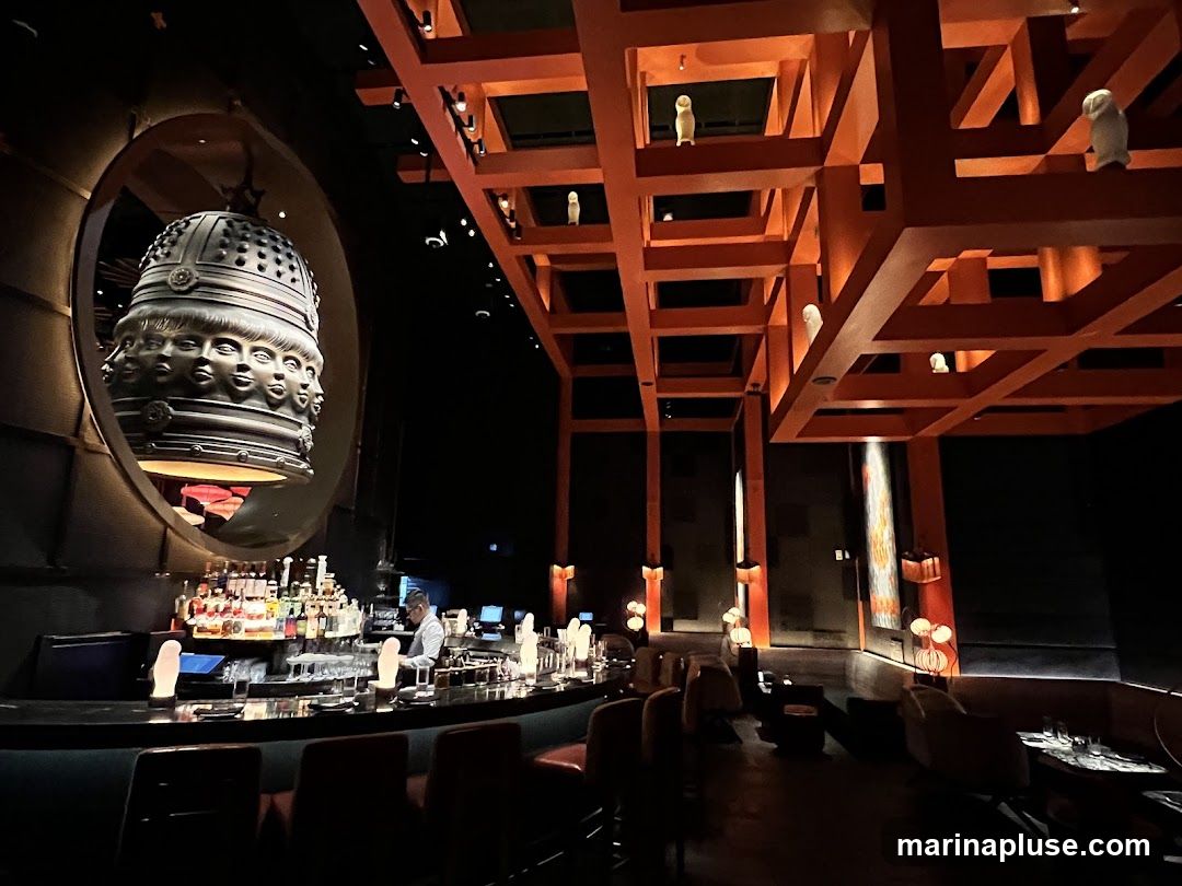 The image shows the interior of a dimly lit bar or restaurant. A prominent circular sculpture with multiple faces hangs from the ceiling. Above, a wooden lattice structure with small figures is visible. A long bar with bottles and glassware is manned by a bartender. Seating areas with tables and chairs are arranged throughout the space.