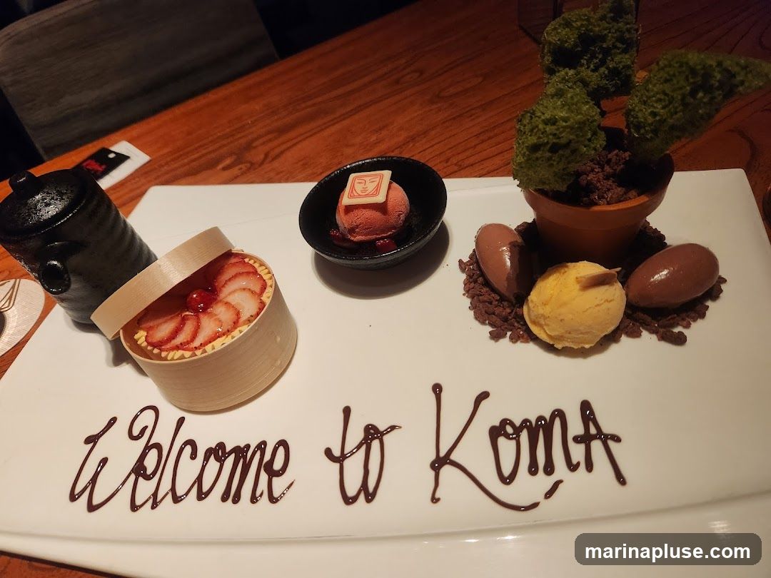 A white platter holds a dessert presentation. In the foreground, chocolate sauce spells out "Welcome to Koma." To the left, a small wooden container holds a strawberry tart. Next to it is a black teapot. In the center, a small bowl contains a scoop of pink sorbet topped with a square of white chocolate. To the right, a terracotta pot with green, moss-like decorations sits on a bed of chocolate crumbles. Surrounding the pot are three scoops of ice cream: two oval-shaped chocolate ones and one round scoop of yellow ice cream.