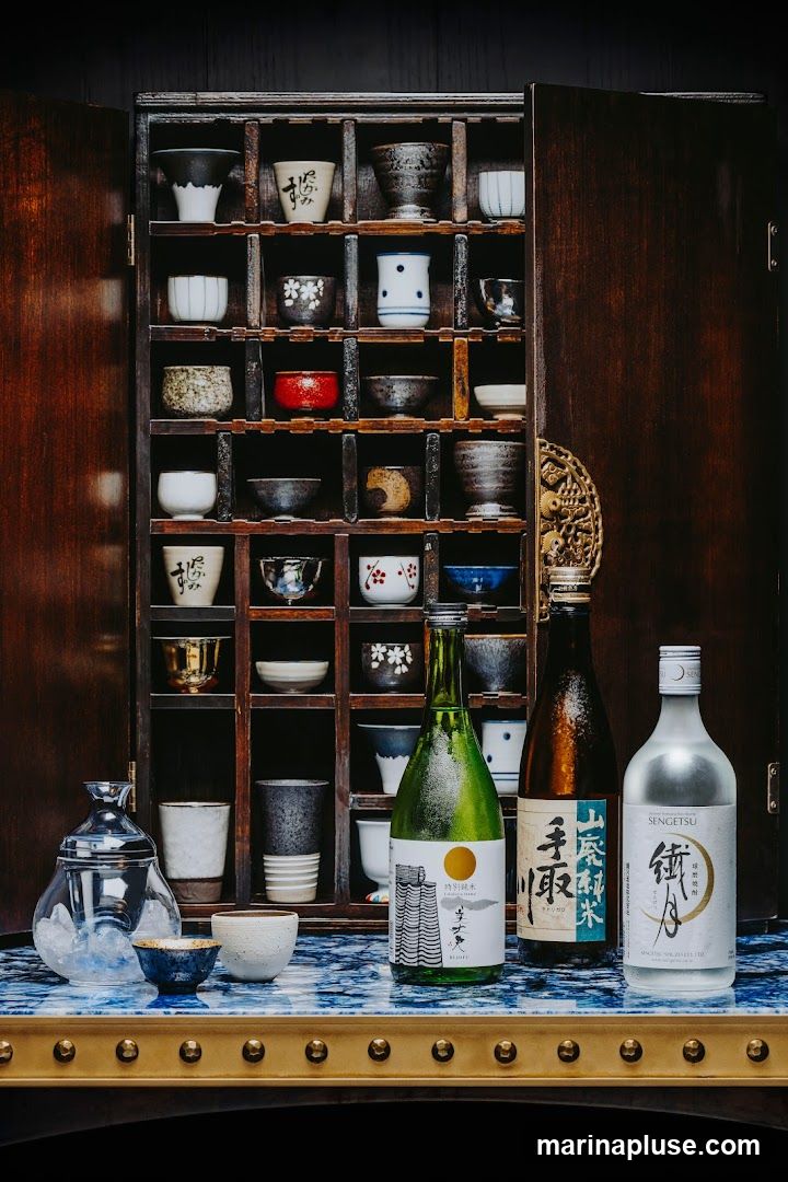 A wooden cabinet filled with various sake cups and bottles. Three bottles of sake are displayed in front of the cabinet on a blue and white patterned surface.