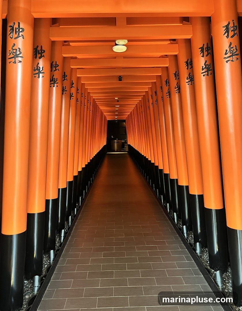A long pathway lined with vibrant orange torii gates, reminiscent of Fushimi Inari Shrine in Kyoto. The gates are adorned with black Japanese calligraphy. The pathway is paved with dark grey rectangular tiles and flanked by black pillars and decorative stones. At the far end of the pathway, a dimly lit reception area with a sign that reads "ROHA" is visible.