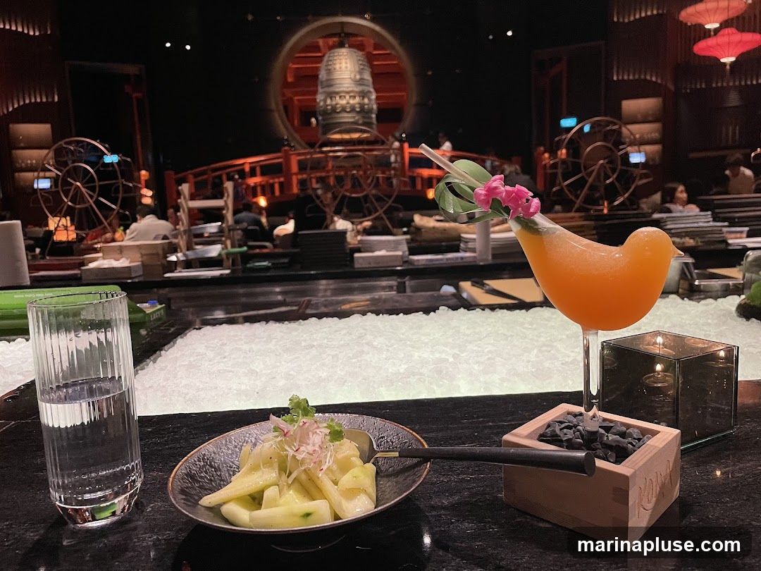 A close-up view of a bar counter with a glass of water, a small appetizer, and a cocktail served in a bird-shaped glass. The background shows a dimly lit restaurant interior with decorative elements and patrons.