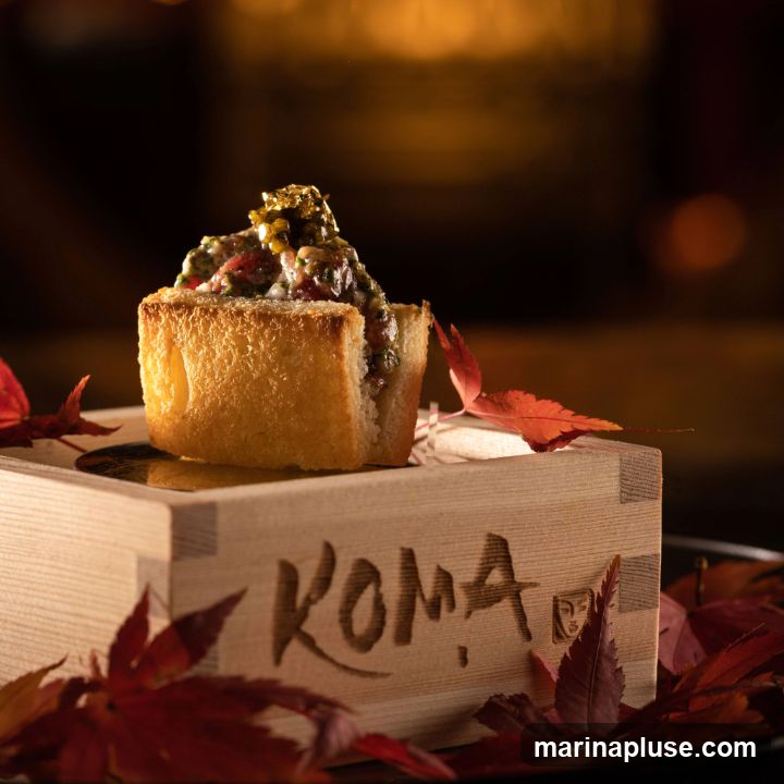 A close-up shot of a gourmet appetizer served on a wooden box. The appetizer consists of a piece of toasted bread topped with a mixture of finely chopped ingredients and garnished with gold leaf. The wooden box has the word "KOMA" carved into its side. Red maple leaves are scattered around the base of the box, suggesting an autumnal theme. The background is softly blurred with warm, golden light.