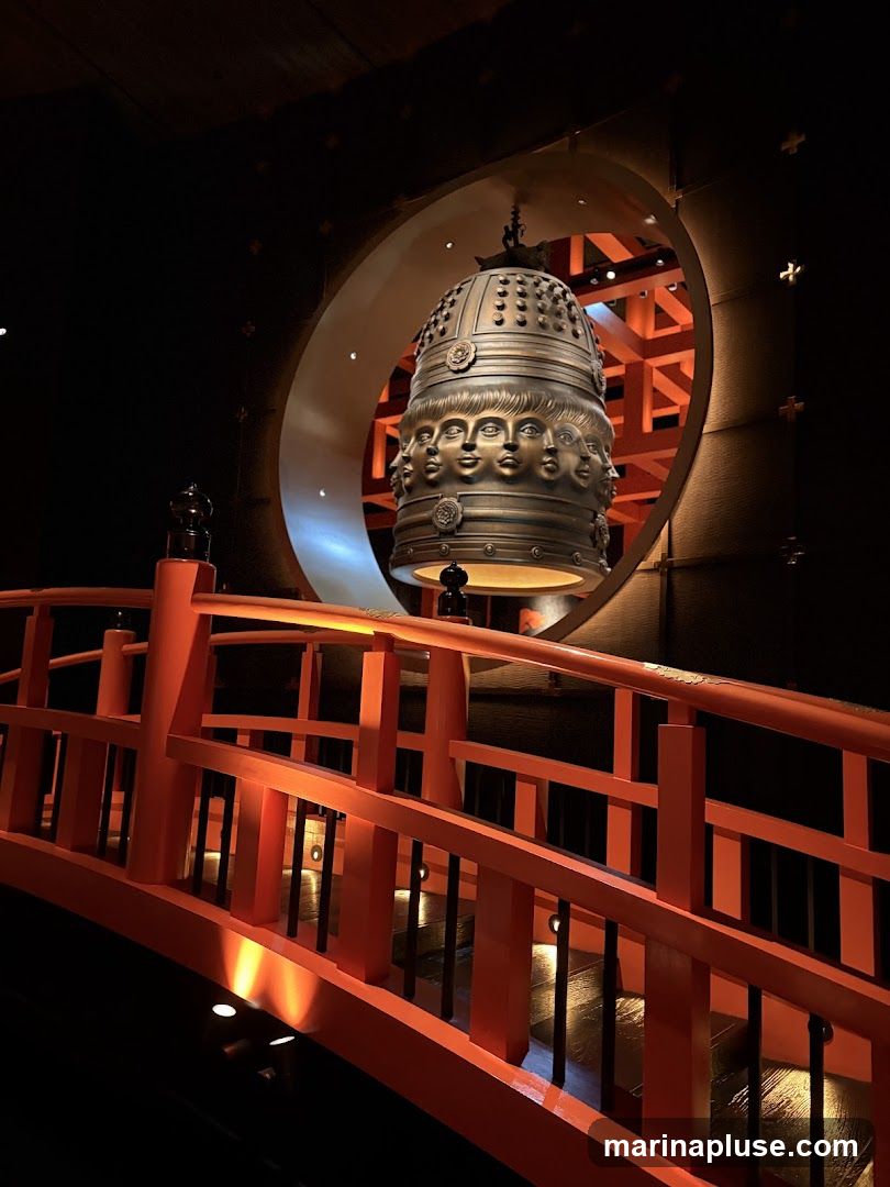 A large, ornate bell adorned with multiple human faces is suspended in front of a circular opening. Below the bell, an orange bridge with black railings curves into the foreground. The lighting is dramatic, casting shadows and highlighting the textures of the bell and bridge.