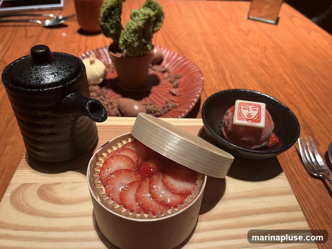 A close-up view of a dessert platter featuring a strawberry tart in a wooden container, a scoop of strawberry ice cream topped with a white chocolate square with a red design, and a small black teapot. In the background, there is a decorative element resembling a cactus in a pot, surrounded by chocolate shavings and small chocolate eggs. The desserts are arranged on a wooden tray on a wooden table.