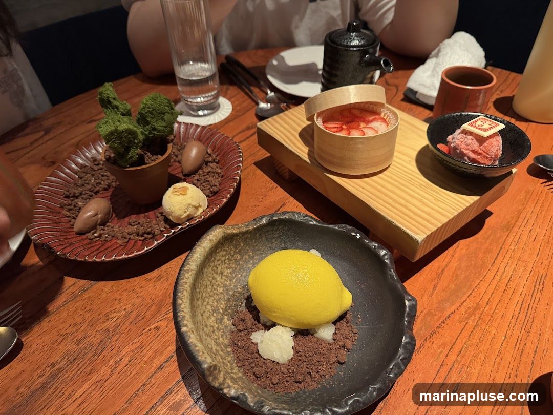 A close-up view of several artfully presented desserts on a wooden table. In the foreground, a dark, textured bowl holds a bright yellow lemon sorbet atop a bed of chocolate crumble and white pieces. To the left, a red, ridged plate features a dessert resembling a potted plant with green foliage, surrounded by chocolate "stones" and a scoop of light-colored ice cream. Further back, a wooden board displays a small wooden container filled with sliced strawberries and a black bowl containing pink sorbet topped with a white chocolate garnish.
