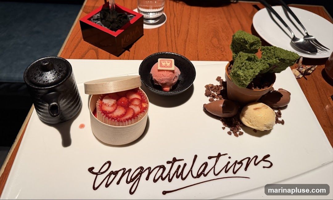A white platter holds a dessert presentation. In the center, a small terracotta pot contains green cake pieces resembling a cactus, surrounded by chocolate crumbs and two chocolate pieces. To the left, a small wooden container is filled with sliced strawberries and a cupcake liner. Next to it is a black teapot. A small bowl contains pink ice cream topped with a white chocolate square. The word "Congratulations" is written in chocolate syrup on the platter.
