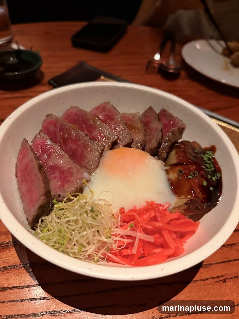 A close-up shot of a donburi bowl filled with sliced beef, a soft-boiled egg, sprouts, and pickled ginger. The dish is served in a white bowl on a wooden table.