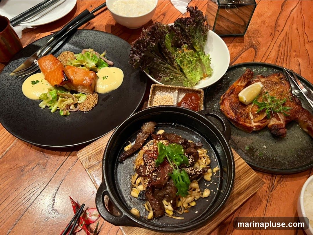 A top-down view of a wooden table laden with several dishes. In the foreground, a cast-iron skillet holds a stir-fry of thinly sliced beef and bean sprouts, garnished with cilantro and sesame seeds. To the left, a black plate features two pieces of glazed salmon served with a creamy yellow sauce and wilted greens. To the right, a dark plate displays a half-roasted chicken, topped with a lemon wedge and fresh herbs. A bowl of red leaf lettuce and a small dish of dipping sauce are positioned behind the chicken. A bowl of white rice and chopsticks are also visible.