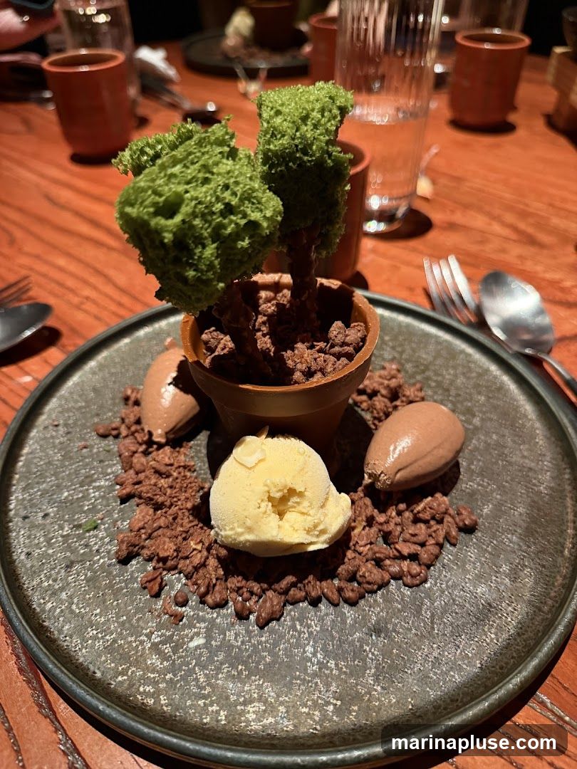 A dessert is presented on a dark plate, resembling a small tree in a pot. The "tree" has green sponge cake foliage and a chocolate trunk, planted in a chocolate-colored pot filled with chocolate crumbles. Around the pot, more chocolate crumbles surround a scoop of yellow ice cream and two chocolate-shaped elements. The setting appears to be a restaurant with wooden tables and blurred background elements including glasses and cups.