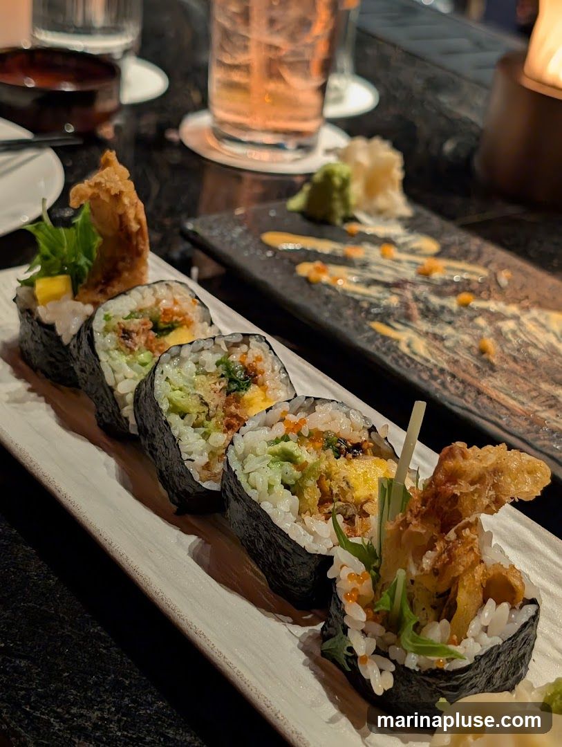 A close-up of several sushi rolls on a white plate. The rolls are made with seaweed, rice, and various fillings, including what appears to be tempura, avocado, and roe. A small dish with wasabi and pickled ginger is visible in the background, along with a drink in a glass.