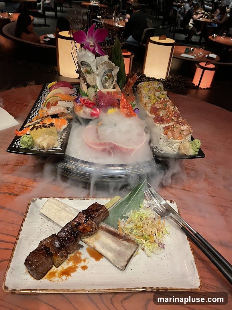 A platter of assorted sushi, sashimi, and oysters is presented with dry ice for a dramatic effect. In the foreground, a skewer of grilled meat sits on a bone, accompanied by a small salad. The setting appears to be a dimly lit restaurant with patrons in the background.