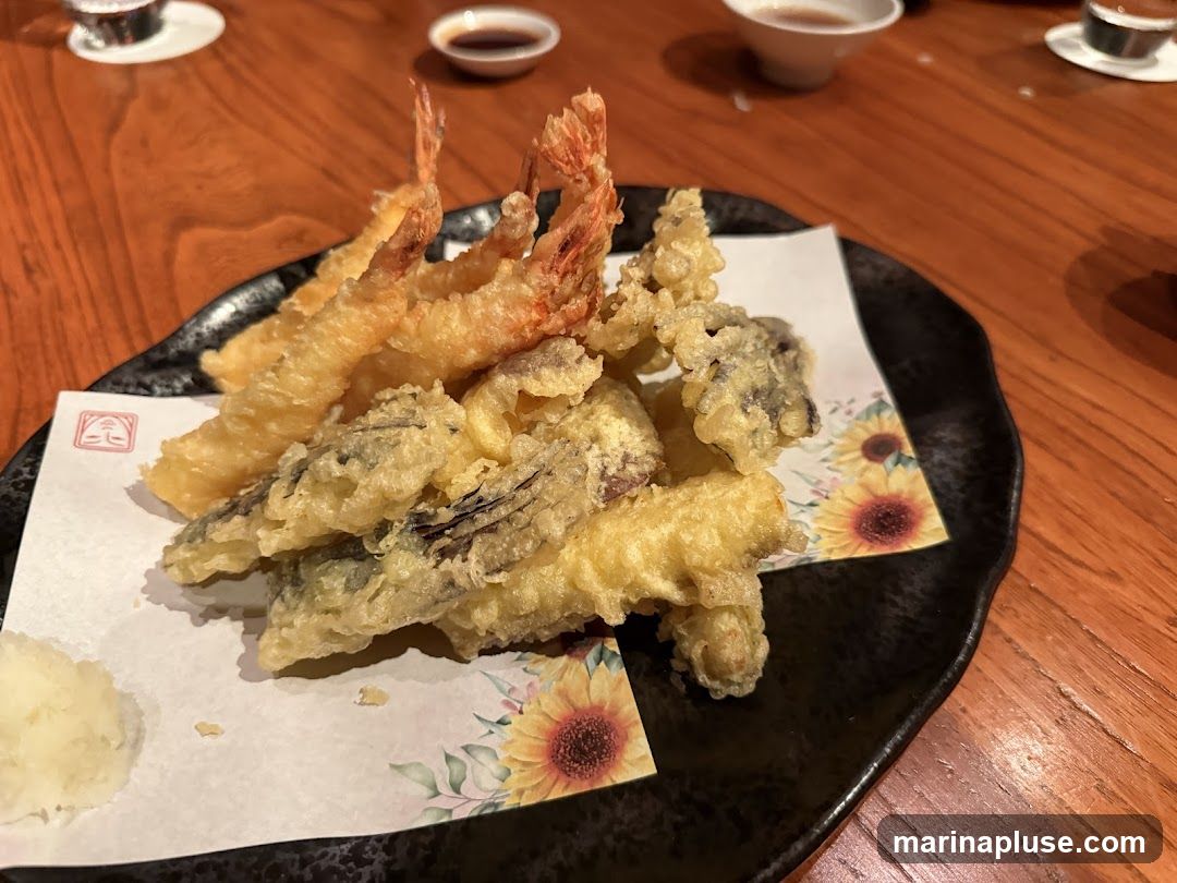 A close-up shot of a tempura platter featuring several pieces of shrimp and what appears to be eggplant tempura, served on a black plate with a decorative paper liner. The paper has a pattern of sunflowers. A small mound of grated daikon radish is on the side. The food is placed on a wooden table, with small dipping bowls visible in the background.