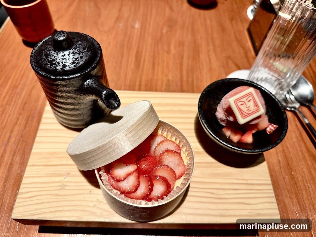 A close-up view of a dessert presentation on a wooden board. In the foreground, a small round container is filled with sliced strawberries arranged over a creamy filling, topped with a few whole strawberries. Next to it, a black bowl holds a scoop of pink ice cream, garnished with a white square chocolate imprinted with a red face design, and surrounded by chopped strawberries. A dark, textured ceramic pitcher with a spout and lid is positioned behind the strawberry dessert. A glass and cutlery are visible in the background.