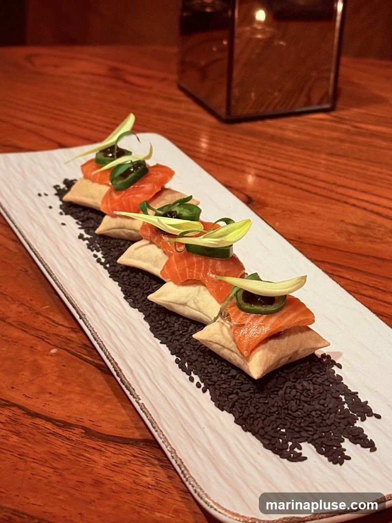 A rectangular white plate with a textured surface holds a row of five salmon bites. Each bite consists of a piece of raw salmon atop a small, square fried pastry. The bites are garnished with thin slices of jalapeño, a drizzle of dark sauce, and a green garnish. The plate is set on a wooden table, and a metallic, reflective cube with a candle inside is visible in the background.