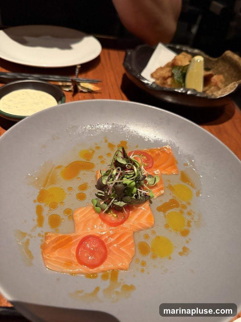 A plate of salmon sashimi is presented with thin slices of cherry tomatoes and a garnish of microgreens. The sashimi is arranged on a grey plate and drizzled with a light dressing. In the background, a small bowl of sauce and a dish of fried food with a lemon wedge are visible.