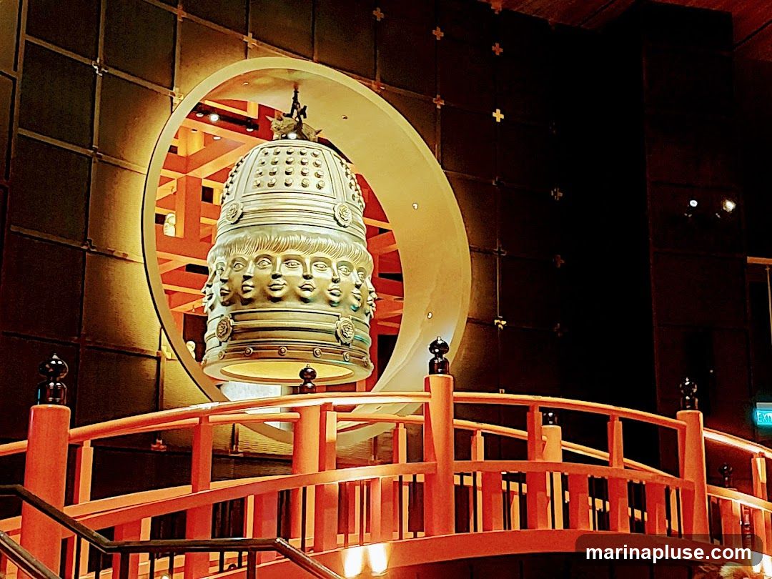 A large, ornate bell adorned with multiple faces hangs in front of a circular architectural feature. In the foreground, a red, arched bridge with railings leads towards the bell. The background features dark, textured walls and illuminated architectural elements.