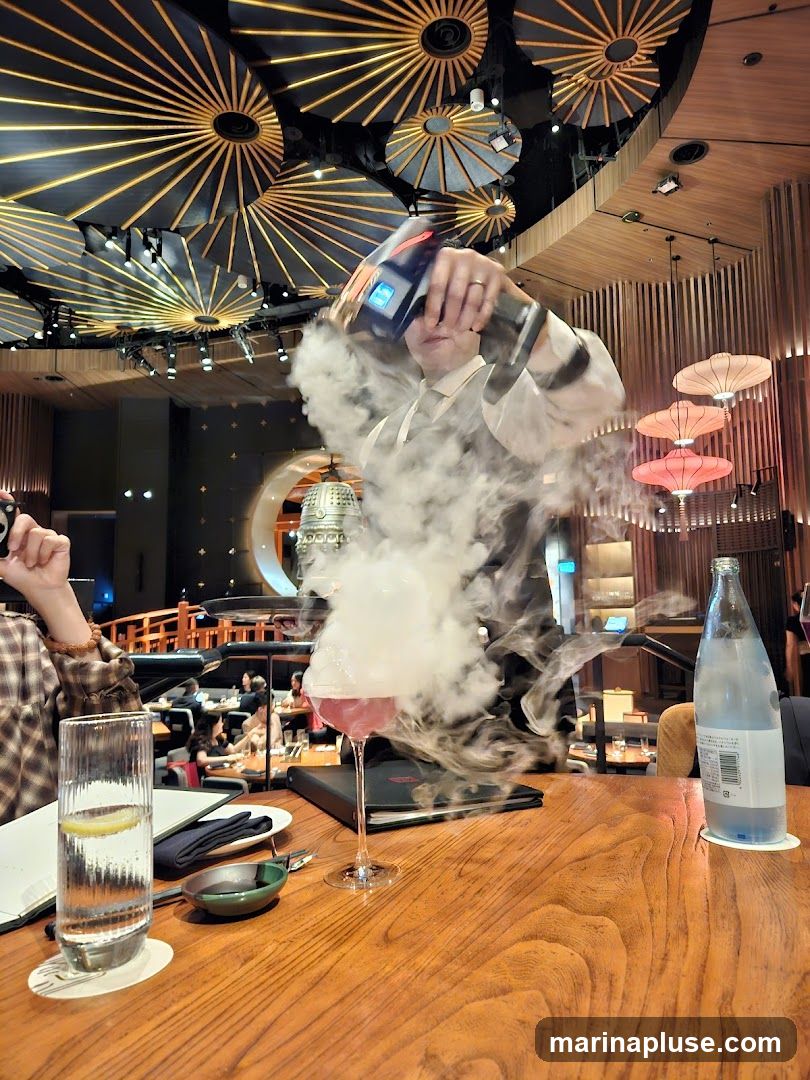 A person is preparing a pink cocktail in a stemmed glass, creating a dramatic smoke effect with a device. The cocktail is on a wooden table in a restaurant with a unique ceiling design featuring radial patterns and lighting. Other elements on the table include a glass of water with lemon, a small dish, and a bottle of water. In the background, other diners are visible, and the restaurant's interior features warm lighting and decorative elements.