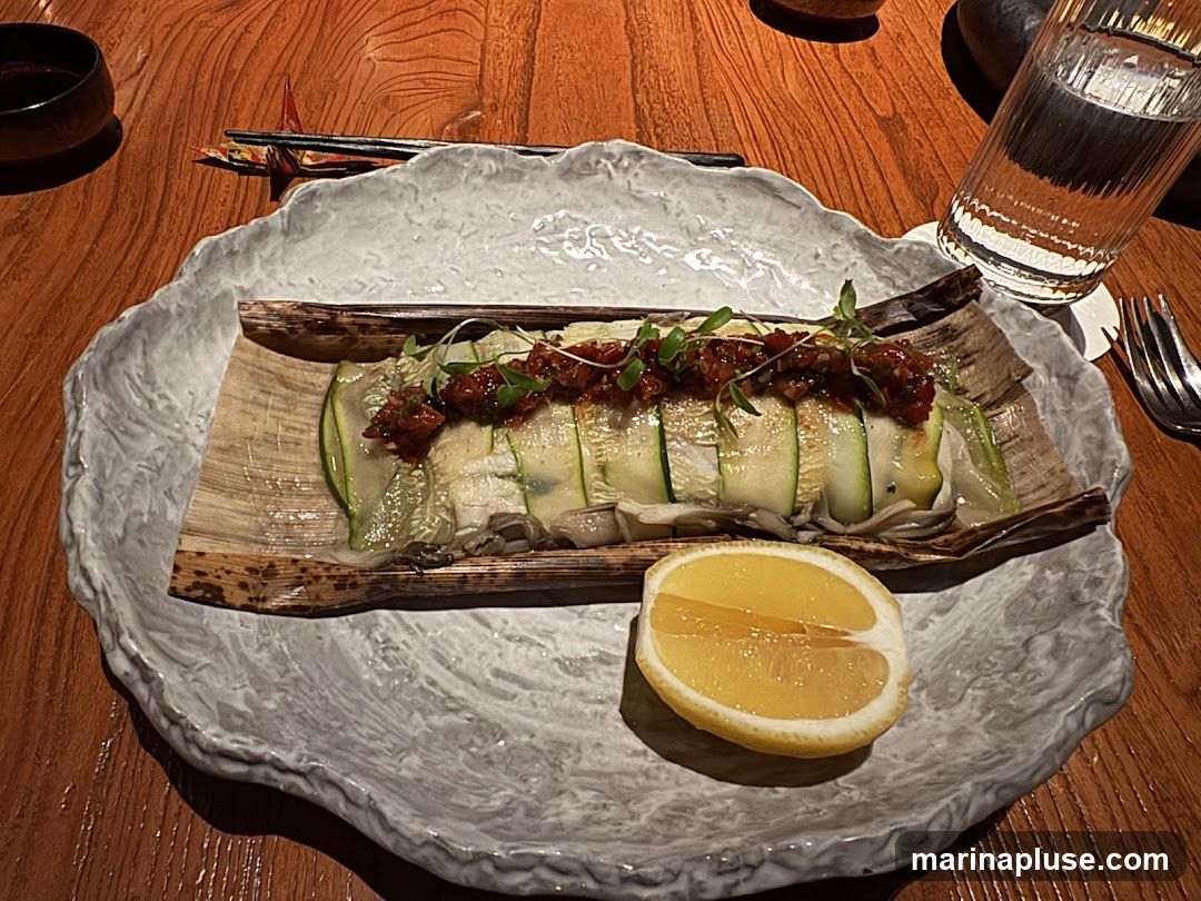 A close-up view of a dish served on a textured, light-colored plate. The food is presented on a bed of what appears to be bamboo leaves, topped with thinly sliced zucchini arranged in a layered fashion. A dark, chunky relish or sauce is spread over the zucchini, garnished with microgreens. A slice of lemon is placed on the side of the plate. The setting includes a wooden table, a small dark bowl, chopsticks, and a glass of water.