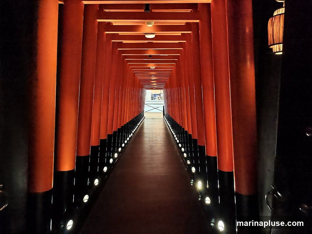A long, narrow pathway is lined on both sides by a repeating pattern of vibrant orange torii gates. The gates are supported by thick, cylindrical pillars that are black at the base and transition to orange. Small, circular lights are embedded in the floor along the base of the pillars, casting a warm glow. The ceiling is also lined with the orange beams of the torii gates, creating a tunnel effect. At the far end of the tunnel, a brighter, more open space is visible, with what appears to be a railing and some architectural elements. A decorative lantern hangs on the right side of the image, near the foreground.