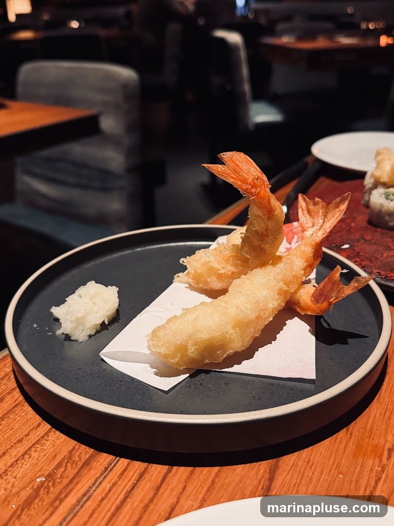 A close-up shot of two pieces of tempura shrimp served on a dark plate with a small mound of grated daikon radish. The shrimp are golden brown and crispy, with their tails still attached. The plate is set on a wooden table in a dimly lit restaurant. In the background, out of focus, are tables and chairs, suggesting a dining environment.