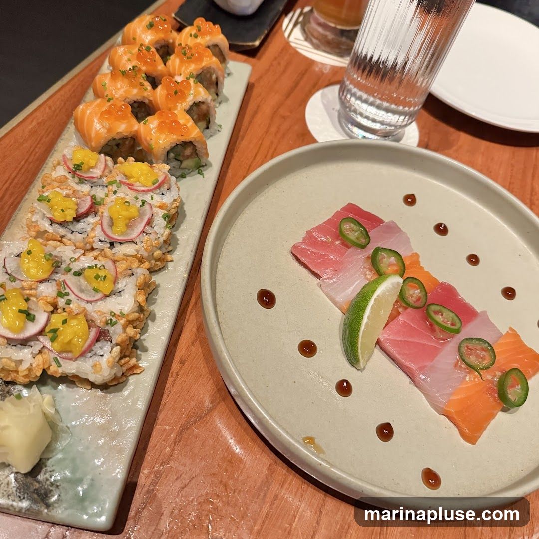 A close-up view of a sushi and sashimi platter on a wooden table. In the foreground, a plate holds slices of tuna, white fish, and salmon sashimi, garnished with jalapeño slices, lime, and a drizzle of sauce. To the left, a long rectangular plate displays two types of sushi rolls. The top roll features salmon with roe, while the bottom roll is topped with radish and a yellow sauce. A small dish of pickled ginger is visible at the bottom left. A glass of water and a white plate are in the background.