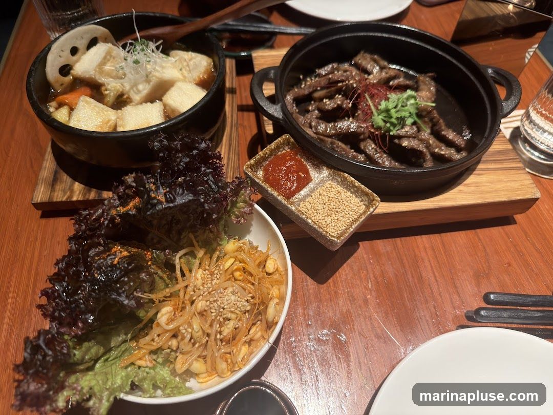 A table set with a Korean BBQ meal. In the foreground, a bowl contains lettuce and seasoned bean sprouts. To the right, a small dish holds two dipping sauces, one red and one sesame seeds. Behind that, a cast-iron pan holds thinly sliced grilled beef, garnished with cilantro and red chili threads. In the background, a dark bowl contains a stew with tofu, lotus root, and vegetables.