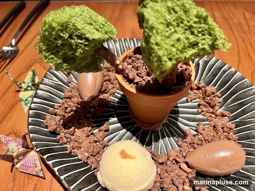 A dessert is presented on a black and white striped plate, designed to look like a small potted plant. The "plant" has a green, airy cake or meringue "foliage" attached to a chocolate "stem" and is planted in a small orange pot filled with chocolate crumble resembling soil. More chocolate crumble is scattered around the plate, along with two quenelles of chocolate mousse and a scoop of vanilla ice cream. Two origami cranes, one green and one pink, are placed on the side of the plate. A fork and knife are visible in the upper left corner.