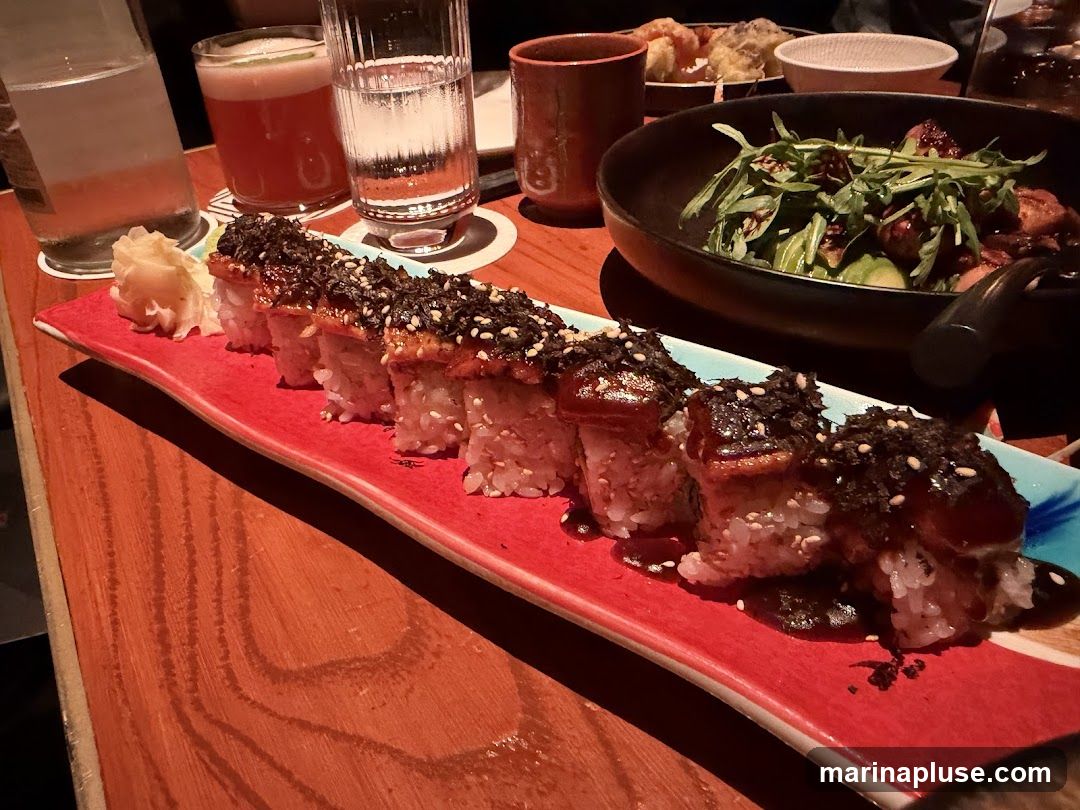 A close-up shot of a sushi roll with eel and seaweed topping, served on a red platter. In the background, a bowl of salad with greens and what appears to be chicken or seafood is visible, along with drinks and other dishes. The food is arranged on a wooden table.