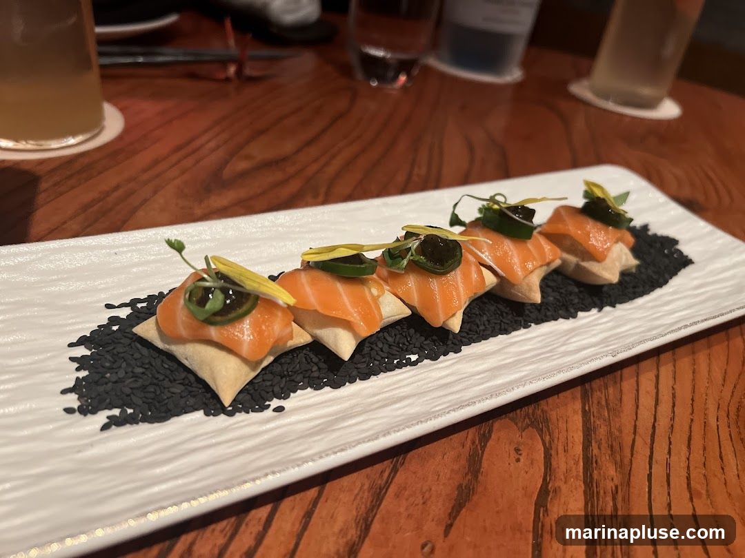 A white rectangular plate holds five salmon bites arranged on a bed of black sesame seeds. Each bite consists of a piece of salmon atop a small, square cracker or pastry, garnished with a slice of jalapeño, microgreens, and a yellow edible flower. The plate is on a wooden table, with drinks and out-of-focus elements in the background.