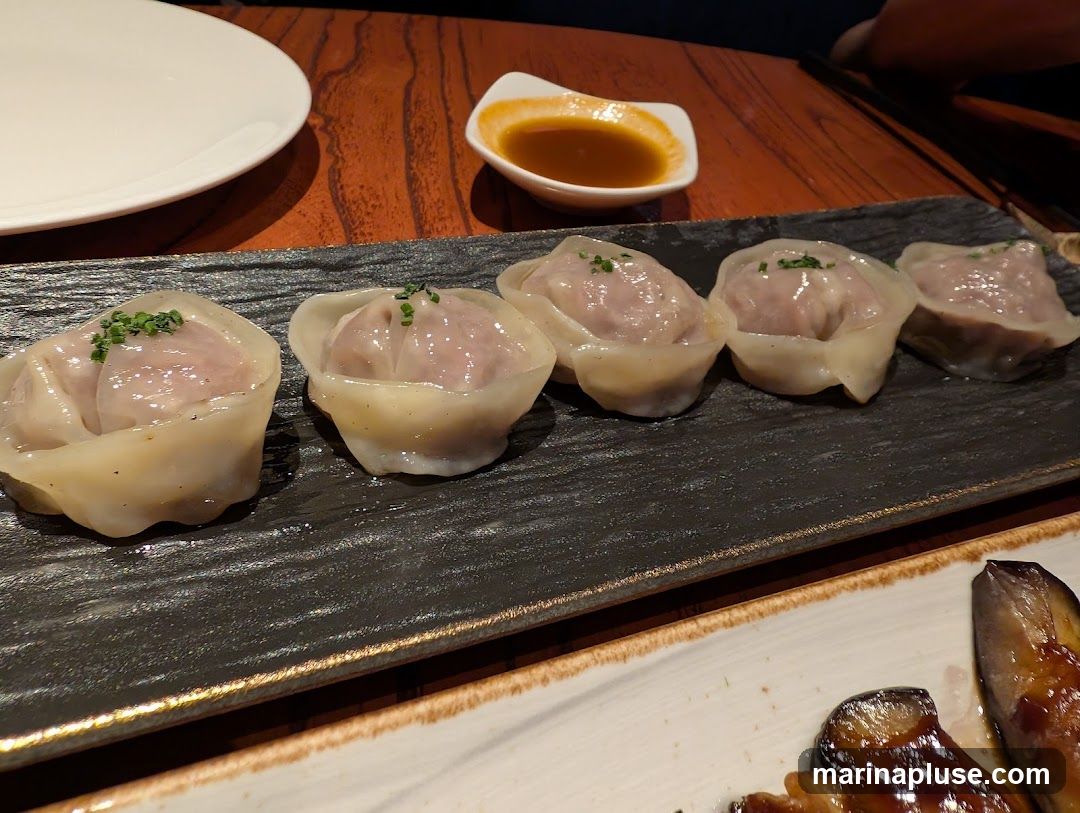 A close-up shot of several steamed dumplings arranged on a dark rectangular plate. The dumplings are filled with a light-colored meat and topped with finely chopped green herbs. A small white dish containing a reddish-brown dipping sauce is visible in the background, along with a white plate and a wooden table surface. Another dish with glazed eggplant is partially visible in the foreground.