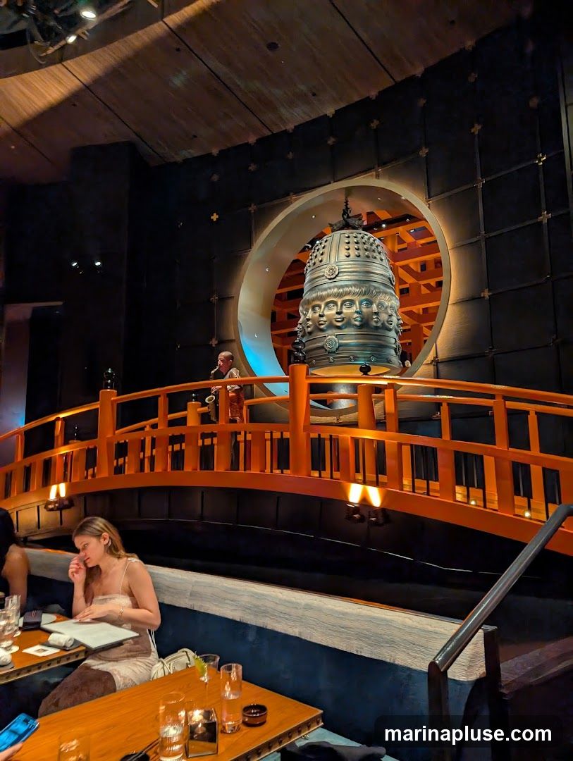 An interior view of a restaurant with a large, ornate bell featuring multiple faces, displayed within a circular opening. An orange bridge with railings spans across the space in front of the bell. A person is visible playing a saxophone on the bridge. Diners are seated at tables in the foreground, with one woman looking at a menu. The ambiance is dimly lit with warm lighting.