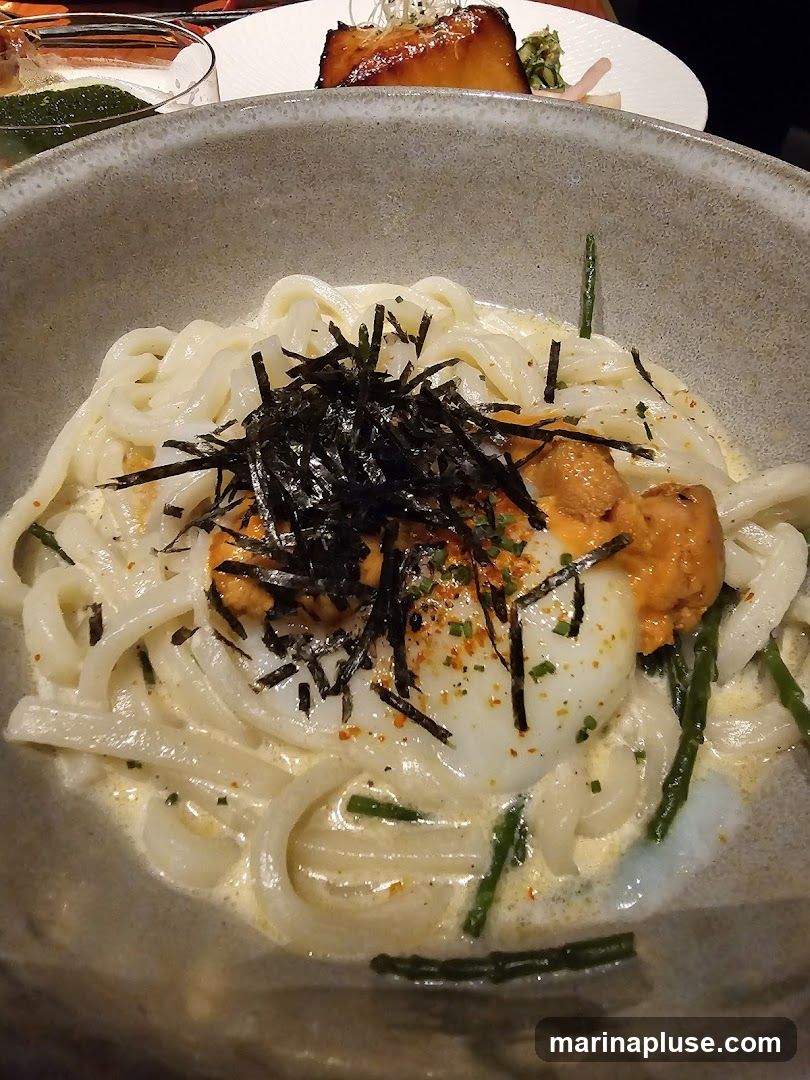 A close-up shot of a bowl of udon noodles in a creamy sauce, topped with sea urchin (uni), a soft-boiled egg, and shredded nori seaweed. Green chives or scallions are also visible. In the background, a piece of grilled fish and a drink are partially visible.