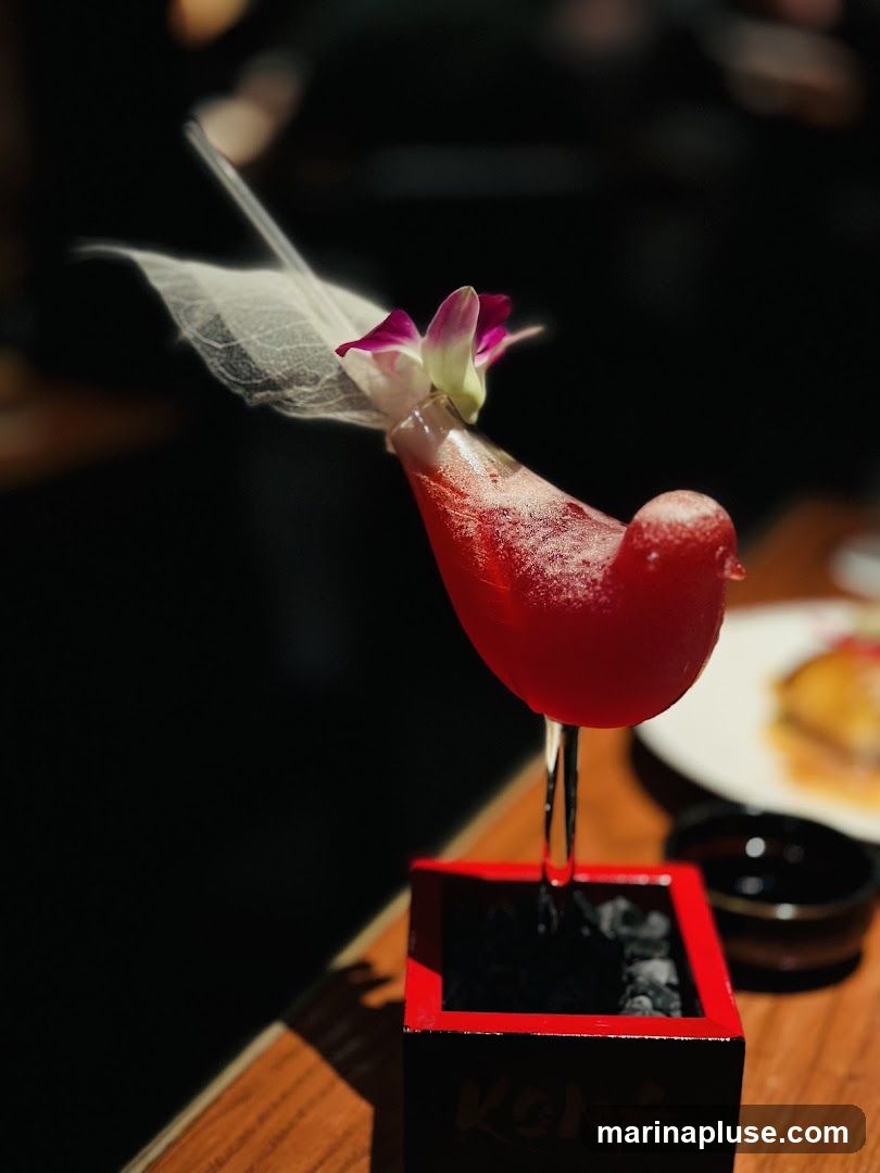 A red cocktail served in a glass shaped like a bird, garnished with a delicate leaf and a purple orchid. The glass is presented in a small red and black square holder with Japanese characters on it. The drink is on a wooden table, with a blurred background suggesting a restaurant setting.