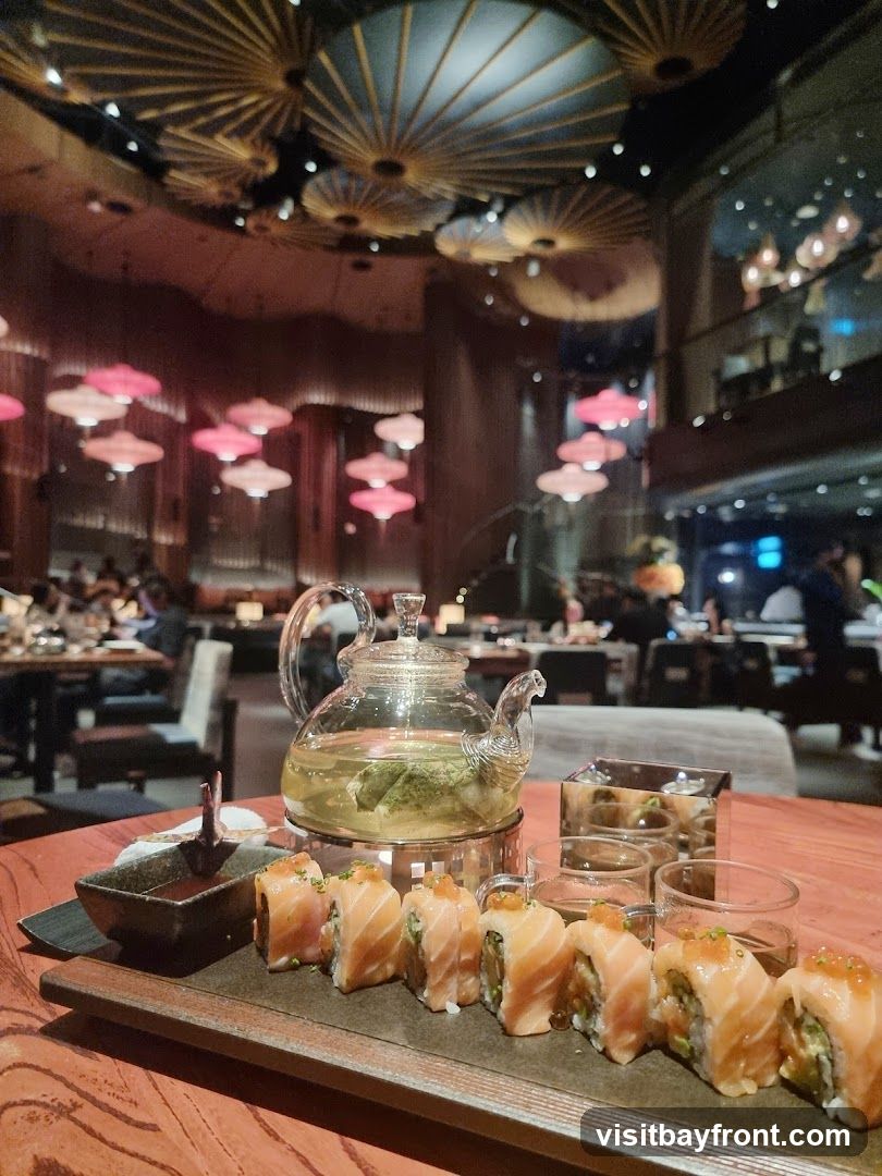 A close-up view of a platter of salmon sushi rolls and a glass teapot filled with green tea, set on a wooden table. In the background, the interior of a restaurant is visible with decorative ceiling lights and patrons dining.