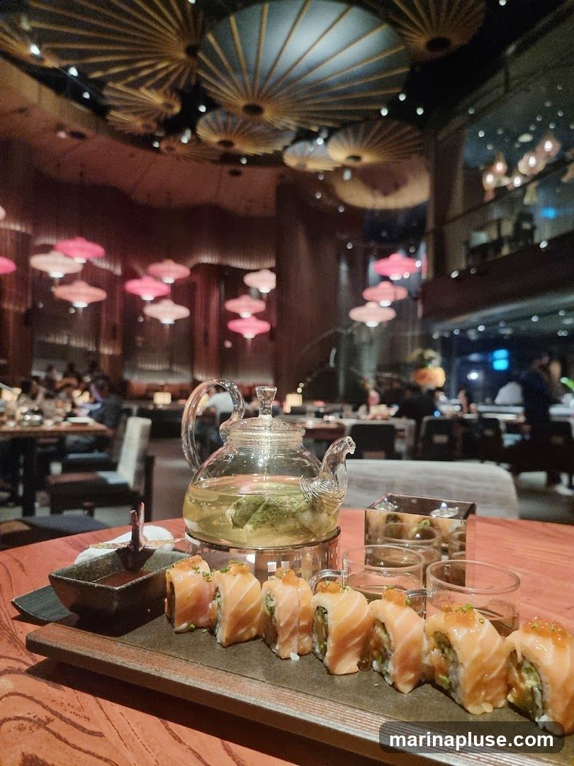 A close-up view of a platter of salmon sushi rolls and a glass teapot filled with green tea, set on a wooden table. In the background, the interior of a restaurant is visible with decorative ceiling lights and patrons dining.