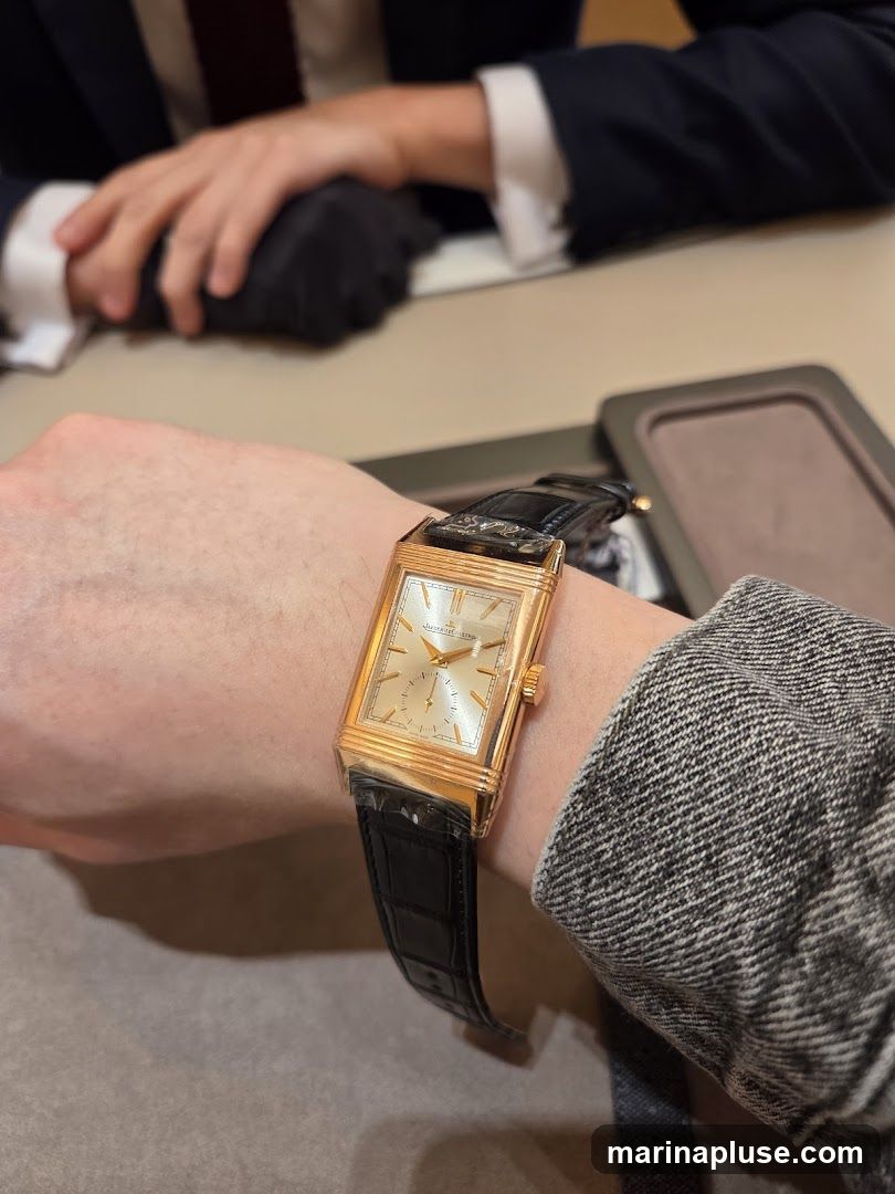 A close-up shot of a person's wrist wearing a rectangular gold watch with a black leather strap. The watch face is silver with gold hands and markers. The watch is on a person's left wrist, and their right arm, wearing a dark suit jacket and white shirt with a burgundy tie, is visible in the background. The setting appears to be a retail environment, possibly a jewelry store.
