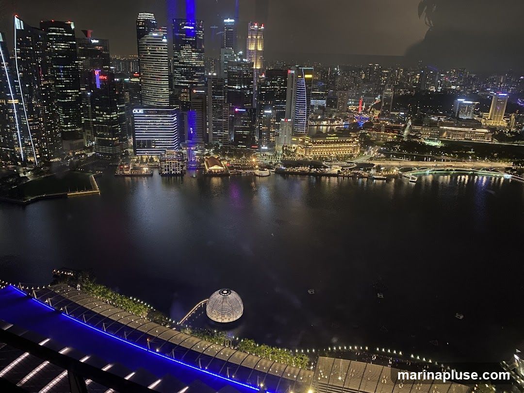 A panoramic view of a city skyline at night, with numerous illuminated skyscrapers reflecting in the dark water of a bay. In the foreground, a modern waterfront promenade with blue lighting and a distinctive dome-shaped structure is visible. The cityscape stretches into the distance, with bridges and more buildings dotting the horizon.