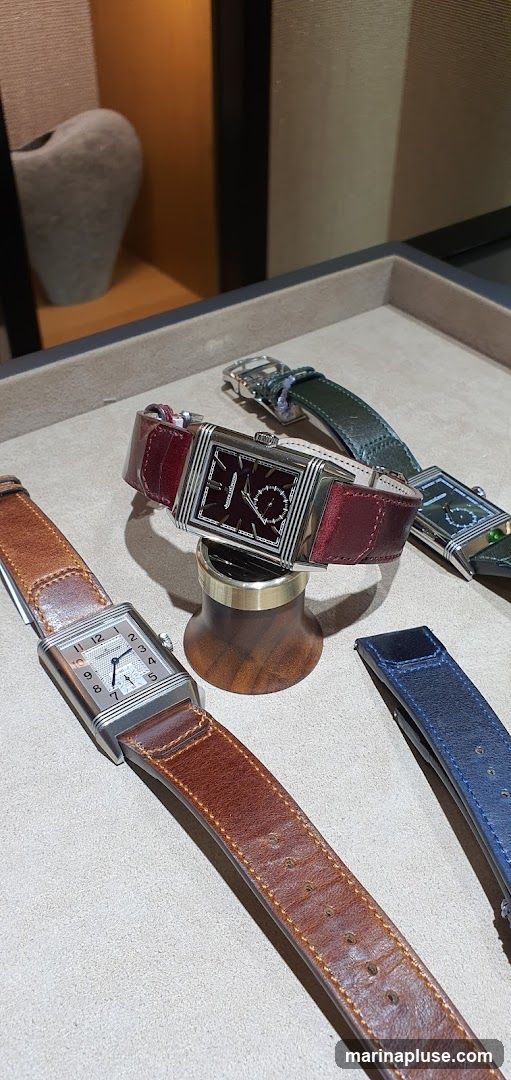 A close-up view of several luxury watches with leather straps displayed on a textured surface. One watch with a burgundy strap is prominently featured on a wooden stand. Another watch with a brown strap is in the foreground, and a watch with a green strap is visible in the background. A blue strap is also visible on the right side. The watches appear to be rectangular with intricate dials.