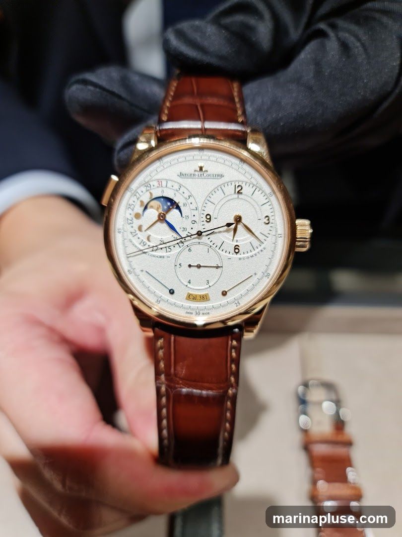 A close-up shot of a Jaeger-LeCoultre watch with a white dial, gold casing, and a brown alligator leather strap. The watch face displays multiple complications, including a moon phase indicator, date, and power reserve. The watch is held by a gloved hand, suggesting it is being presented or displayed. Another watch strap is visible in the background.