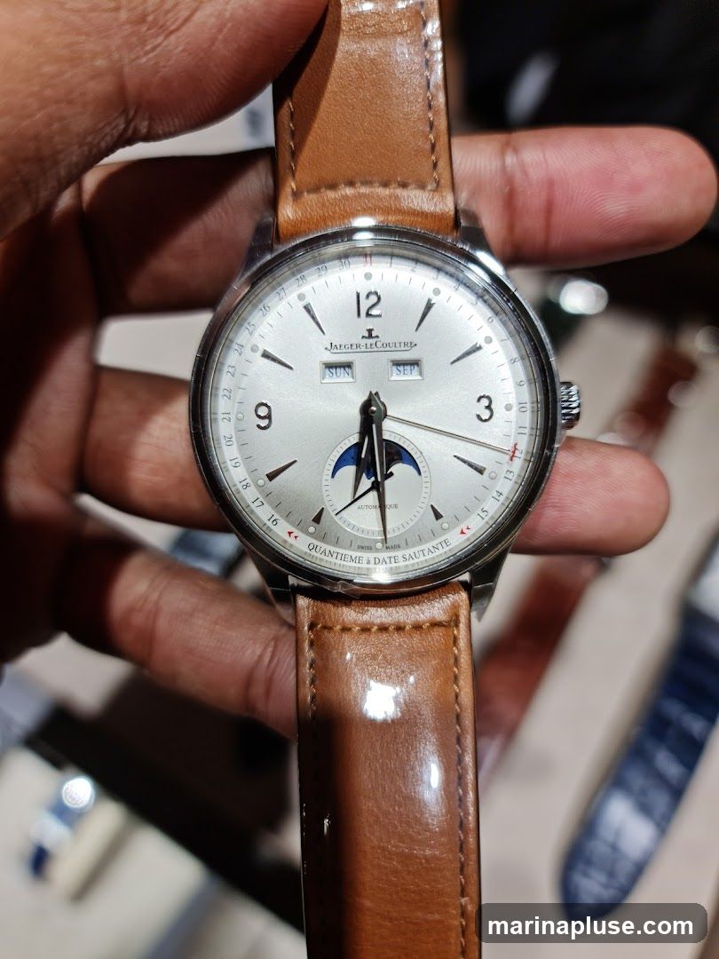 A close-up shot of a Jaeger-LeCoultre watch with a silver dial and a brown leather strap. The watch face displays the time, day, month, and a moon phase complication. The brand name "JAEGER-LECOULTRE" is visible at the top of the dial. The text "QUANTIEME & DATE SAUTANTE" and "AUTOMATIQUE" are also visible.