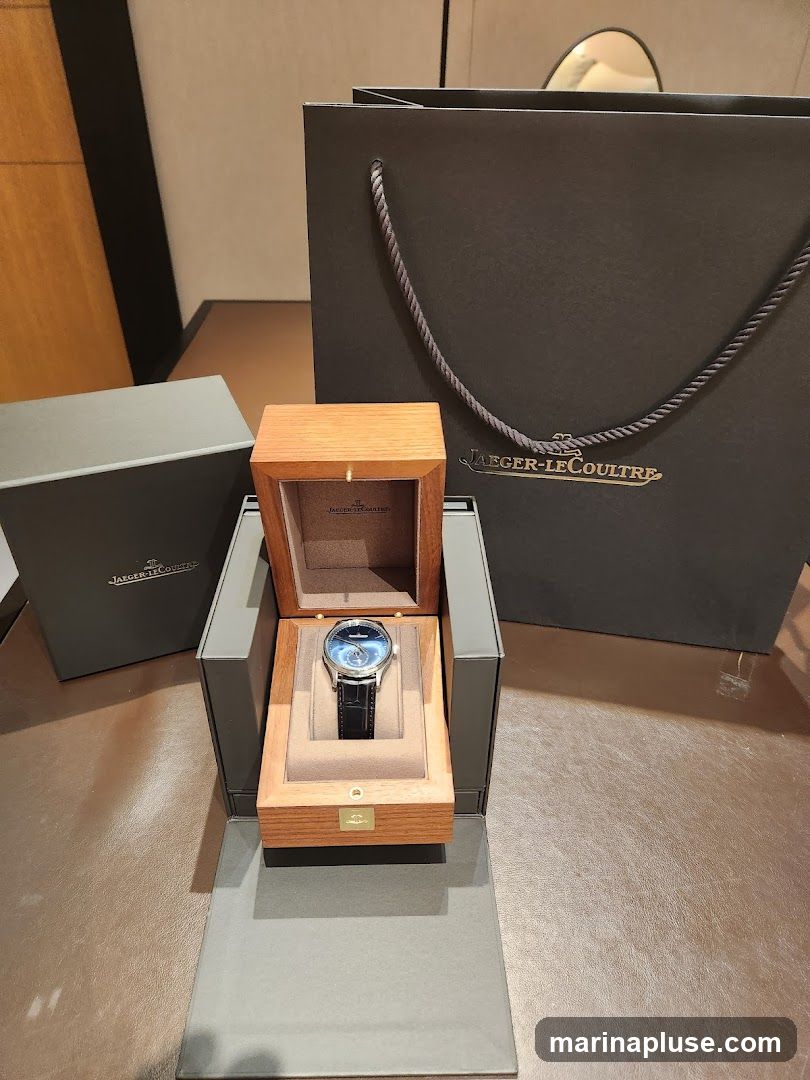A luxury watch with a blue dial and black leather strap is displayed in an open wooden box. The box is part of a larger presentation set, with a grey outer box and a dark grey shopping bag with the Jaeger-LeCoultre logo visible in the background.