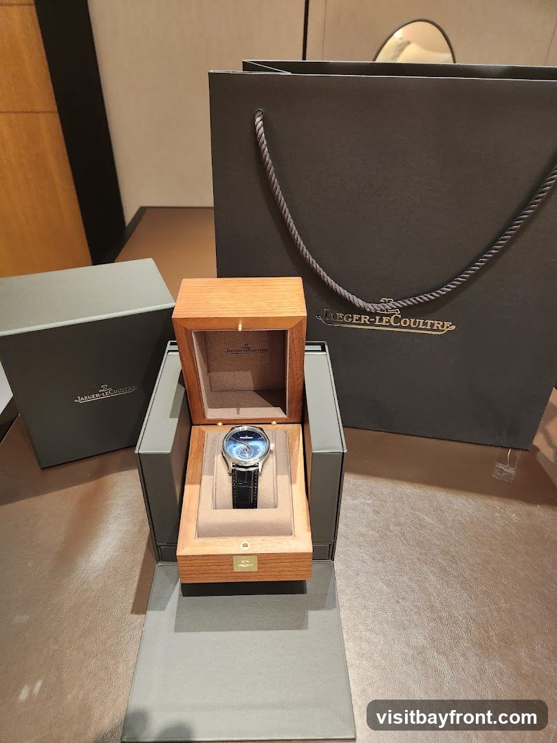 A luxury watch with a blue dial and black leather strap is displayed in an open wooden box. The box is part of a larger presentation set, with a grey outer box and a dark grey shopping bag with the Jaeger-LeCoultre logo visible in the background.
