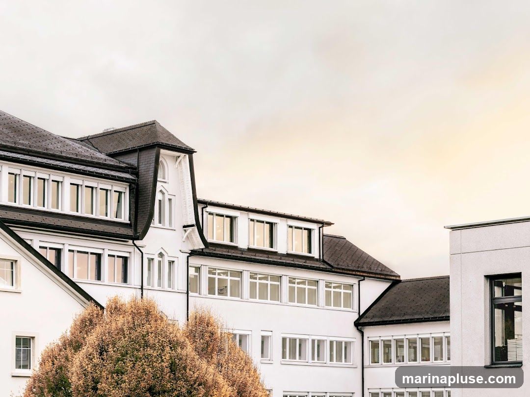 A view of a modern white building with a dark roof and numerous windows. Autumn foliage is visible in the foreground. The sky is overcast with a warm glow.