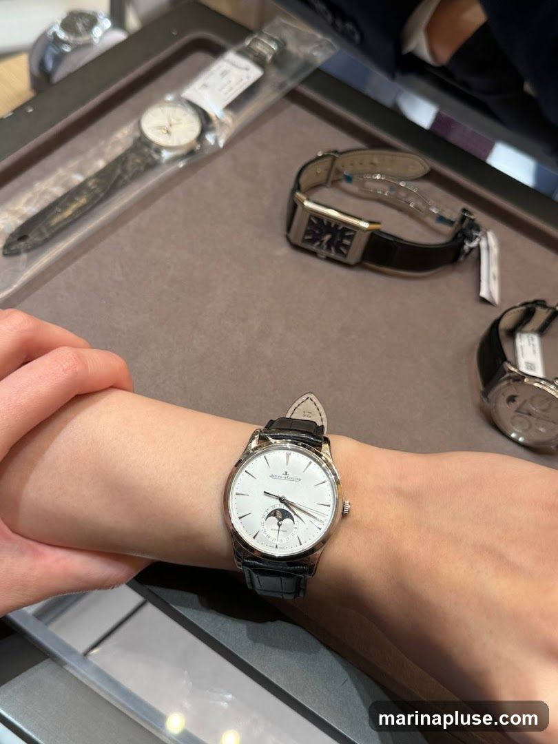 A person's arm wearing a white-faced wristwatch with a black leather strap. The watch has a moon phase complication. In the background, other watches are visible on a display surface, including a rectangular watch with a black strap and a round watch partially visible on the right.