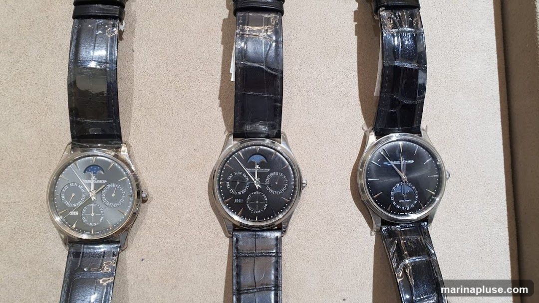 Three luxury watches with black leather straps are displayed side-by-side. The watches have silver casings and feature complex dials with moon phase indicators, day, date, and month complications. The dials are in shades of grey and black.