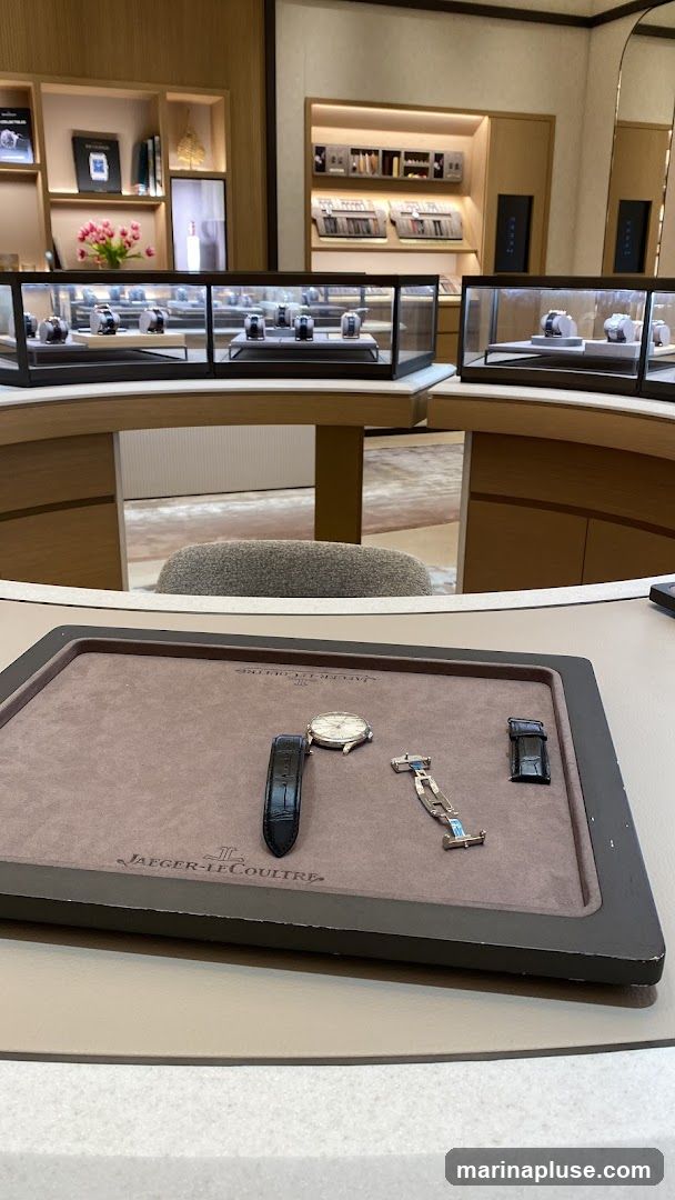 A display tray with a watch and its accessories, including straps and a buckle, is in the foreground. The tray has the Jaeger-LeCoultre logo. In the background, a luxury watch store interior is visible with display cases showcasing watches and shelves with watch-related items.