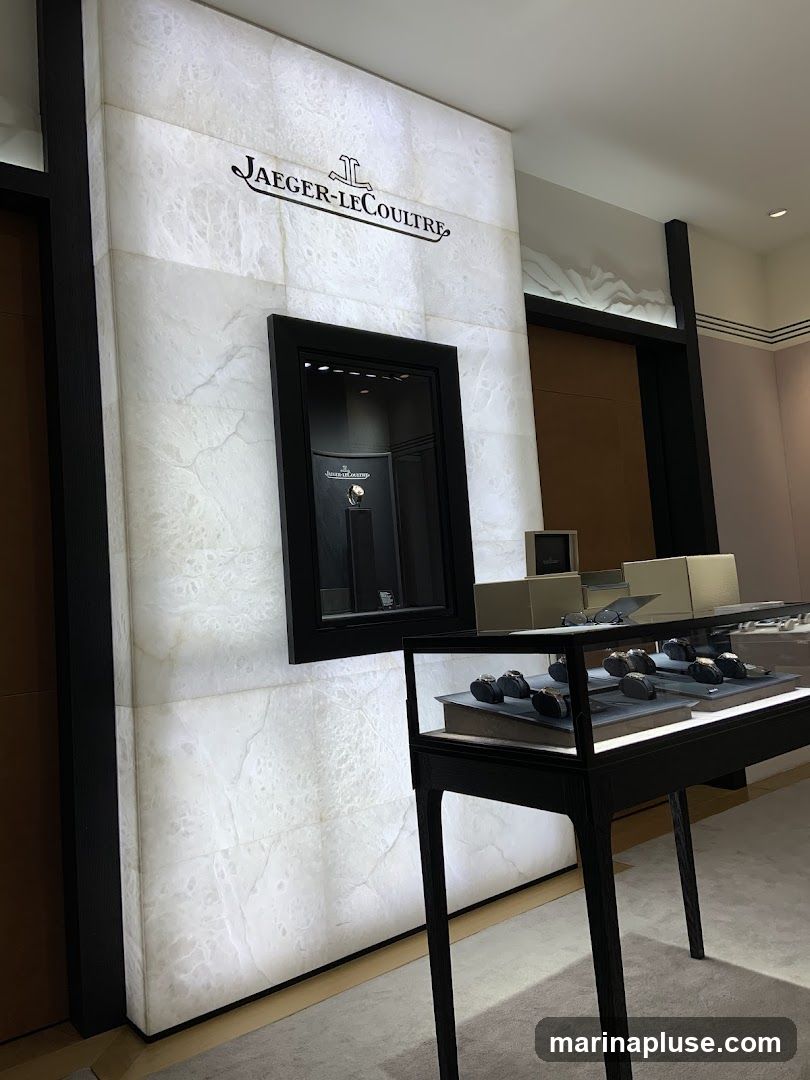 A well-lit interior display featuring the Jaeger-LeCoultre logo prominently on a white, marble-like wall. Below the logo, a recessed display case shows a single watch. To the right, a glass-top display cabinet showcases several luxury watches arranged on stands. The overall ambiance is one of high-end retail.