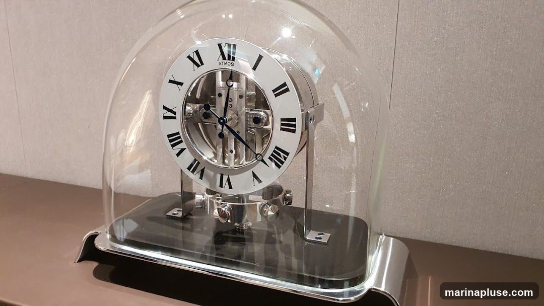 A close-up of an Atmos clock with a glass dome. The clock face features Roman numerals and the brand name "ATMOS" is visible at the top. The intricate mechanical workings of the clock are visible through the face. The clock is displayed on a dark surface with a metallic base.