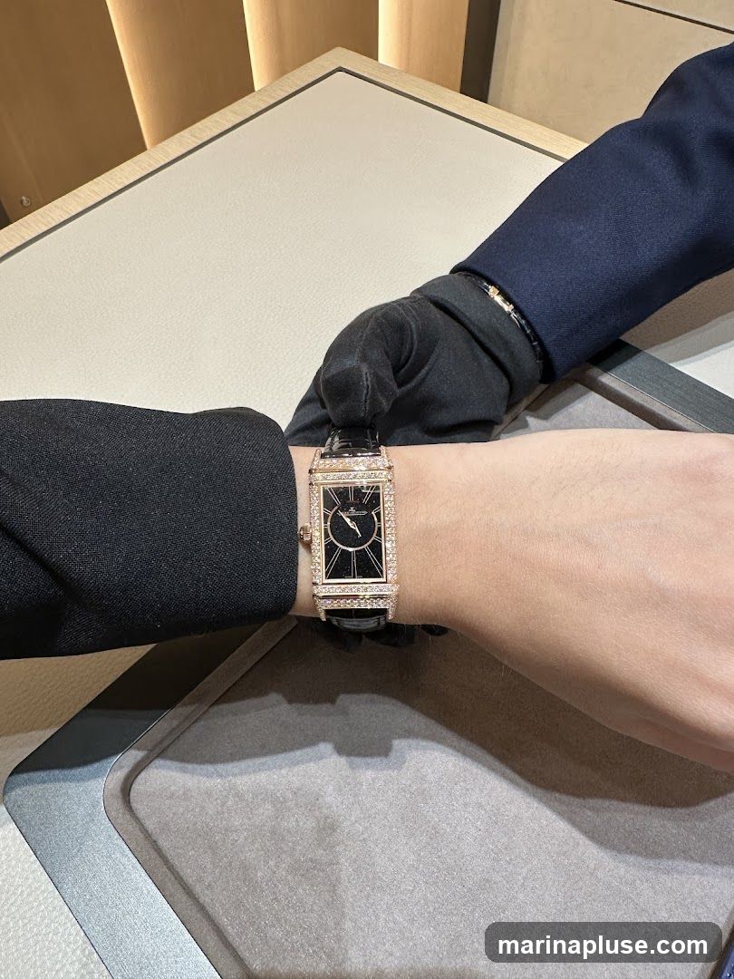A close-up shot shows a rectangular watch with a black face and a diamond-encrusted bezel on a person's wrist. The watch has a black leather strap. Another person, wearing black gloves and a dark suit jacket, is holding the wrist, possibly a salesperson assisting a customer. The background appears to be a display counter in a retail setting.