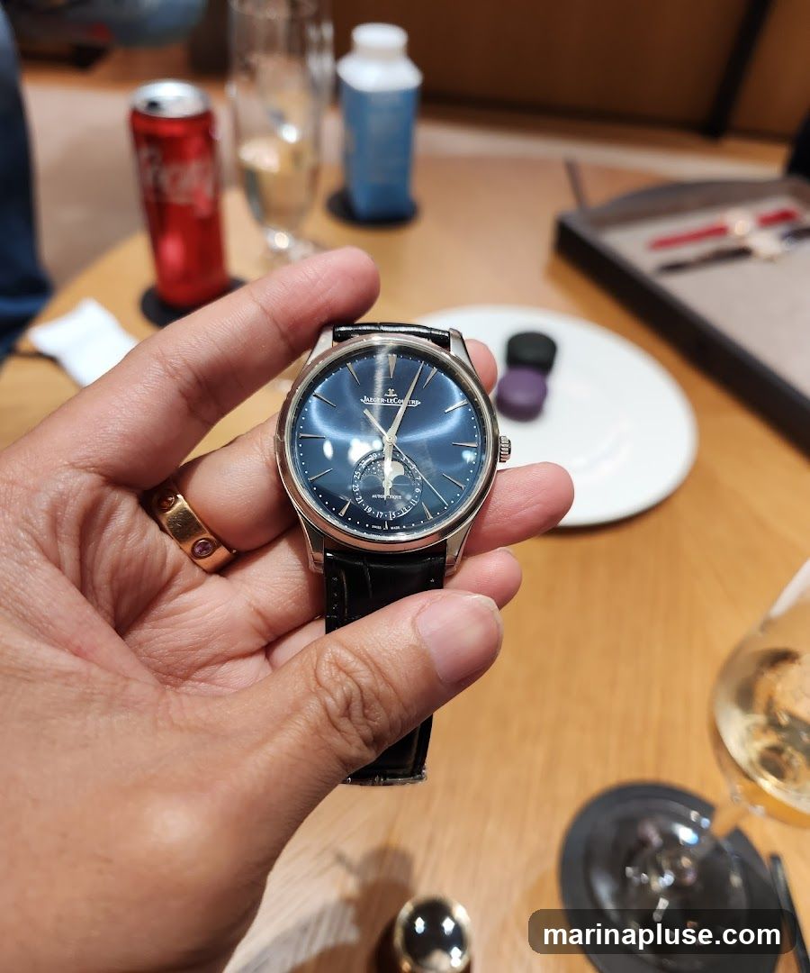 A person is holding a luxury watch with a blue dial and a moon phase complication. The watch has a black leather strap and a silver case. The person is wearing a gold ring with pink stones. In the background, there is a red can of soda, a glass of champagne, a white bottle, a white plate with two small dark objects, and a tray with pens. The setting appears to be a table in a lounge or restaurant.